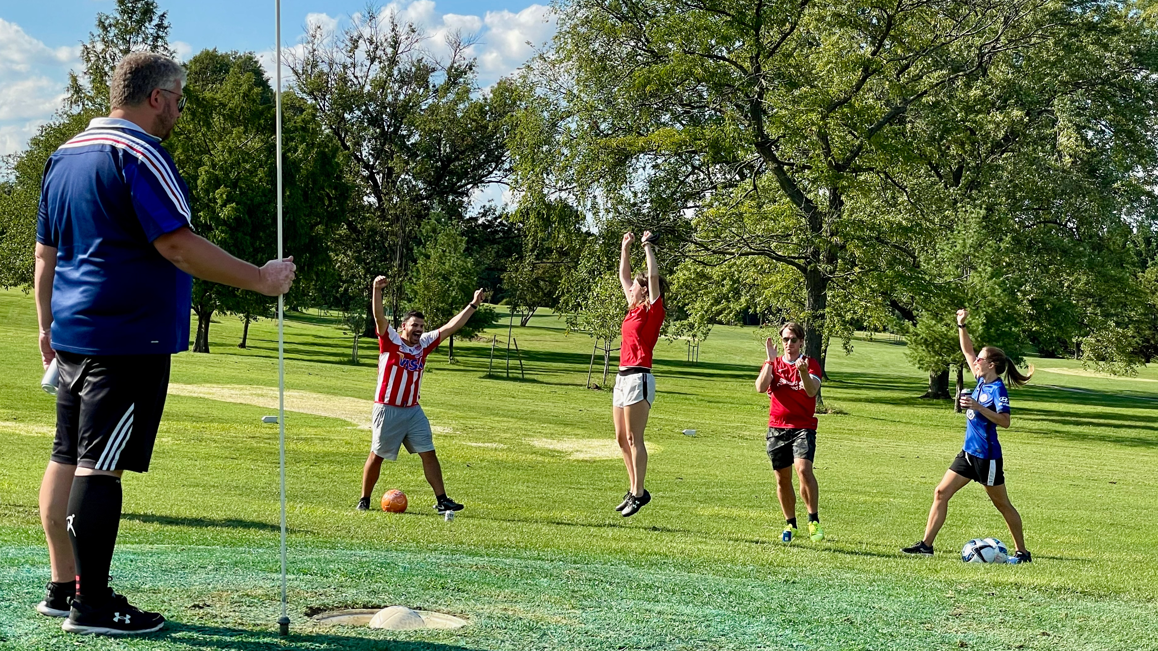 A foursome of footgolf players are excitedly raising hands to cheer as a putted soccer ball sinks in the hole. A course assistant is living the hole flag marker out of the way.